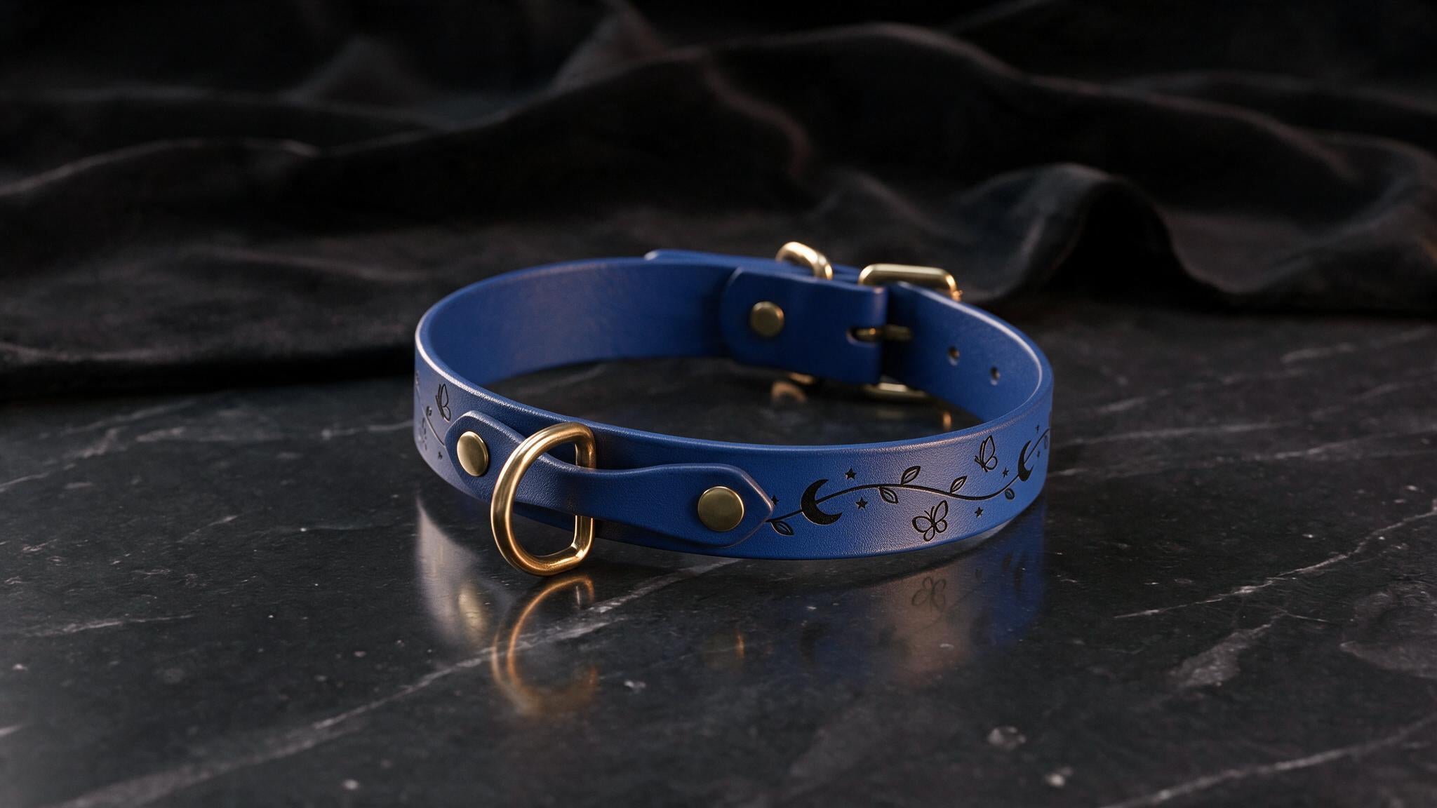 A blue leather collar with a floral design and gold hardware is displayed on a black marble surface.