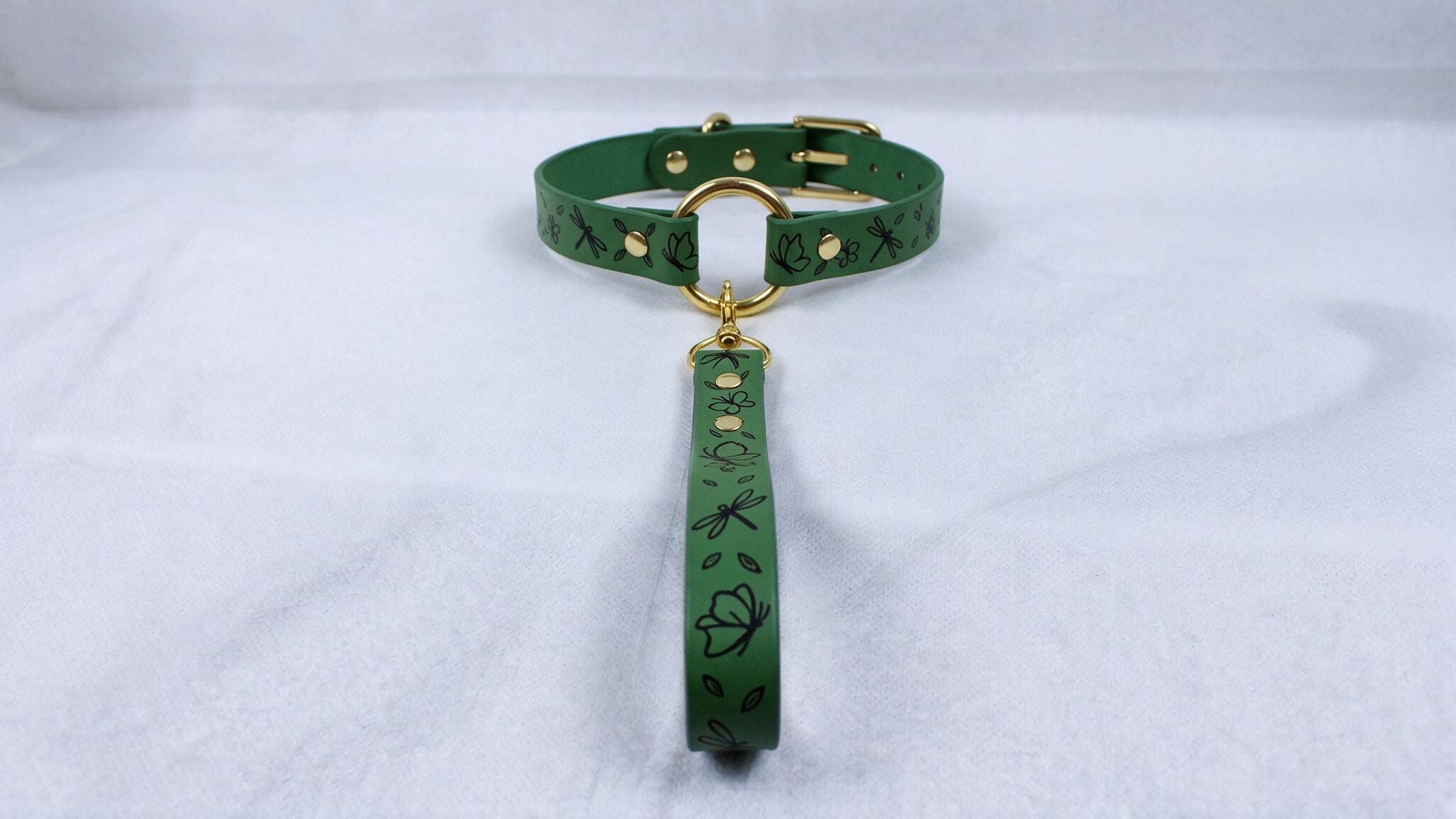 A green collar with a gold buckle and a handle attached to it, placed on a white surface.