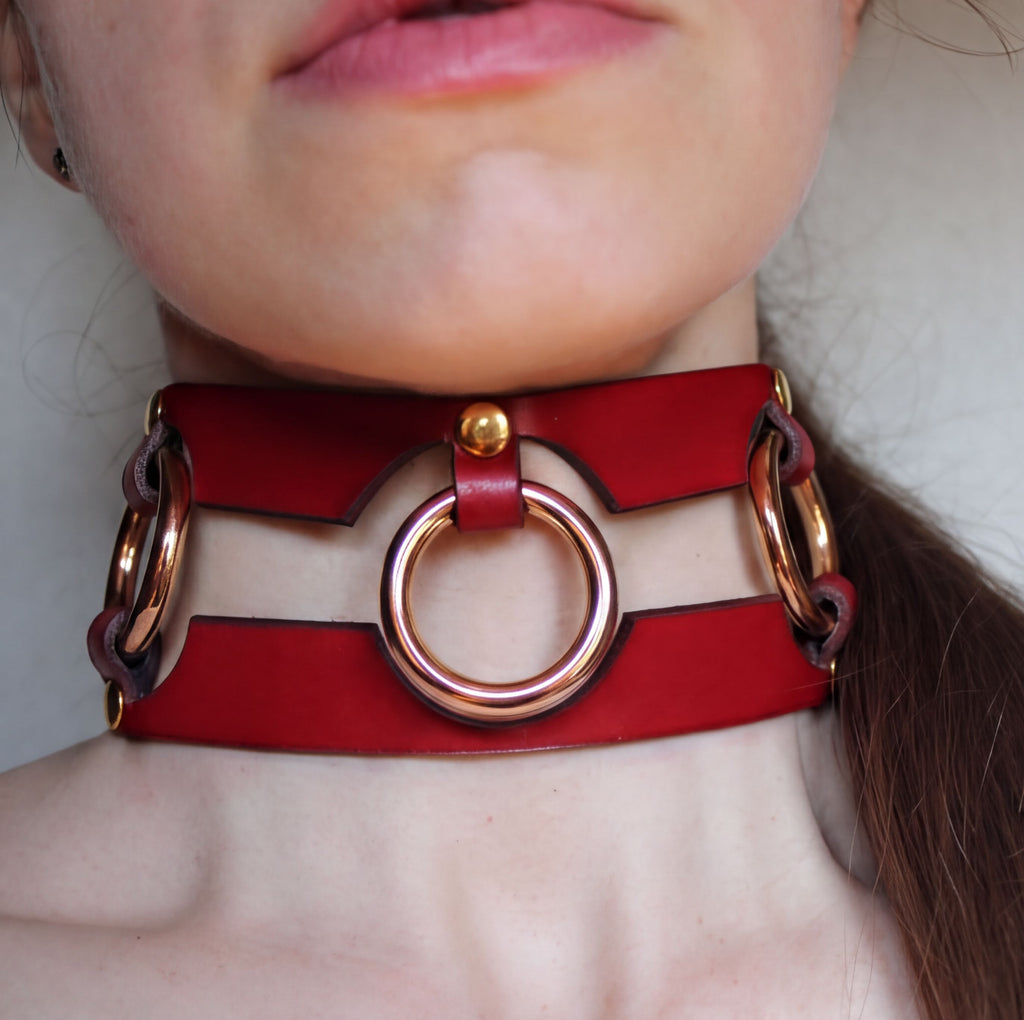 A close-up of a person's neck wearing a red leather choker necklace with a gold-colored ring.