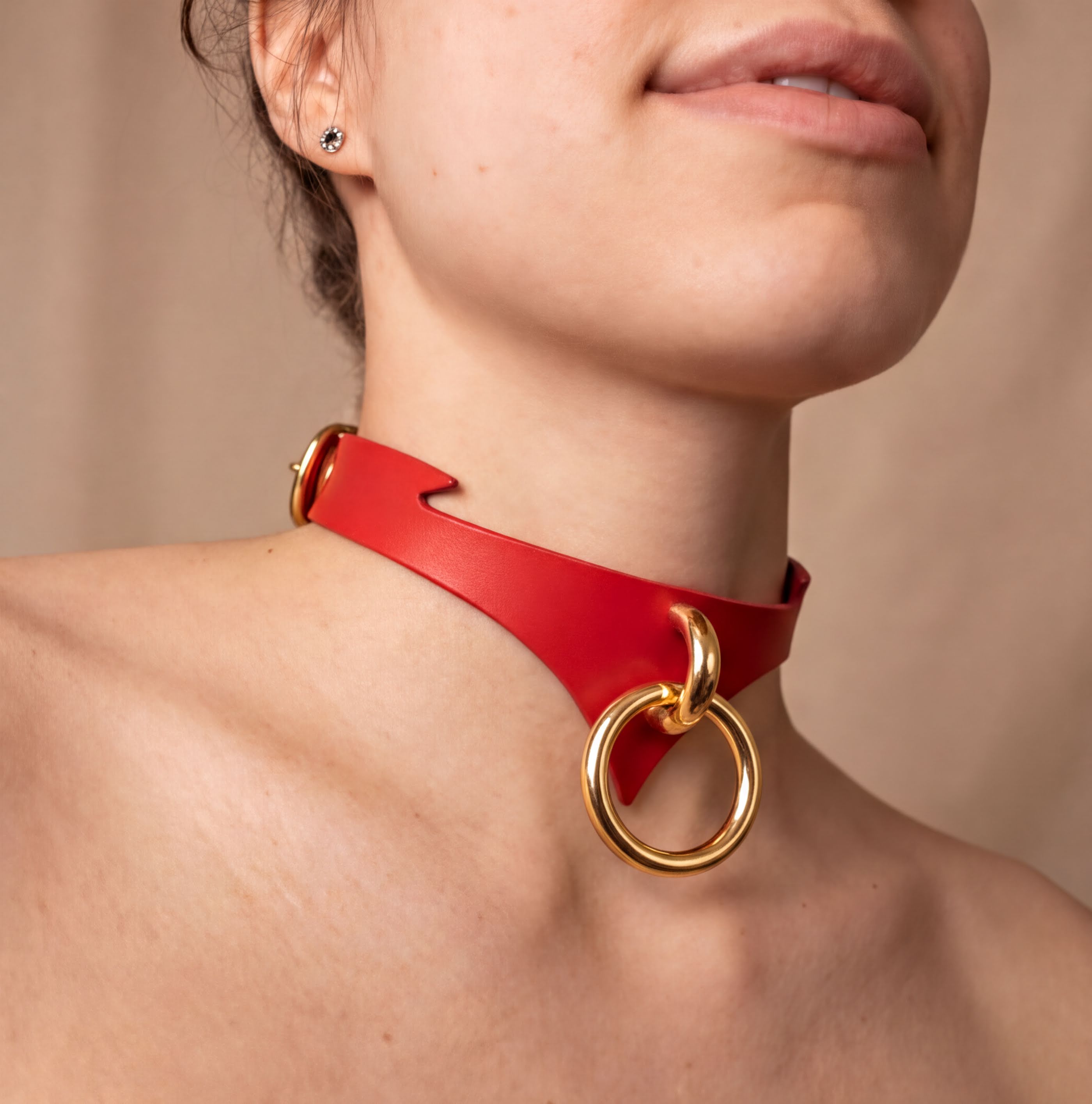 A close-up of a woman's neck and chest area wearing a red leather choker necklace with gold-colored rings.