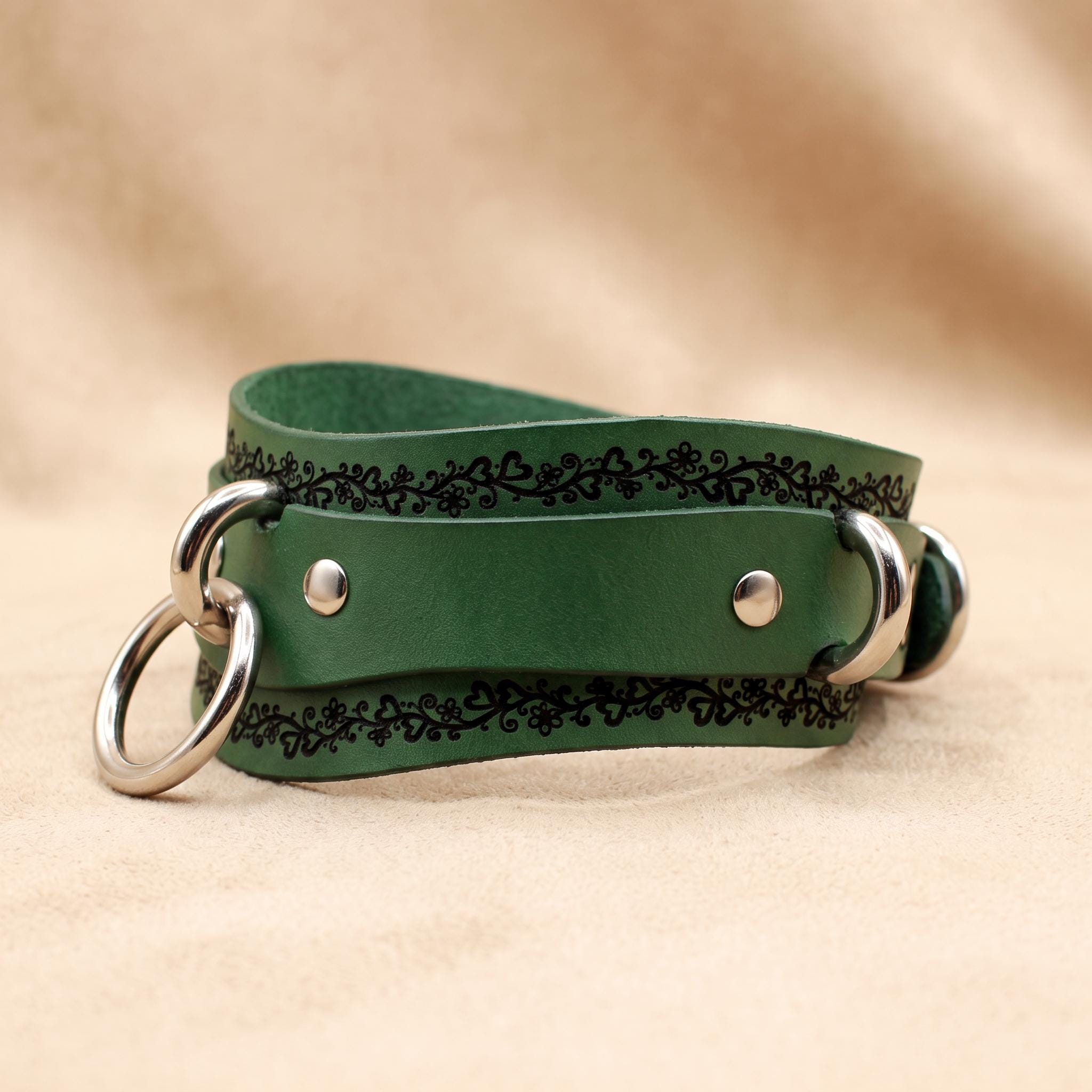 A green leather strap with metal rings and a decorative pattern on the edges, placed on a beige background.