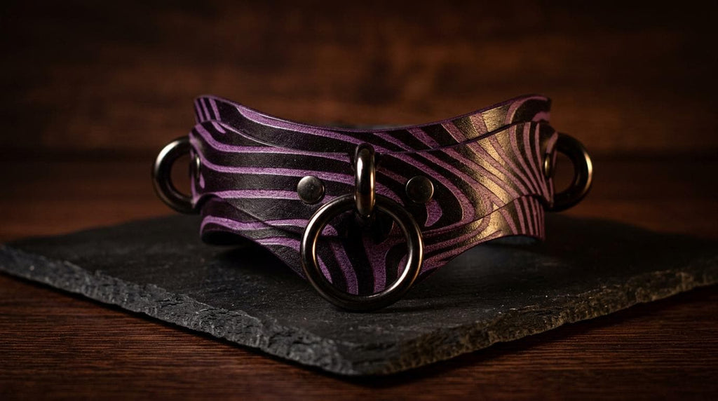 A purple and black zebra-striped leather collar with metal rings and a circular clasp is displayed on a dark surface.