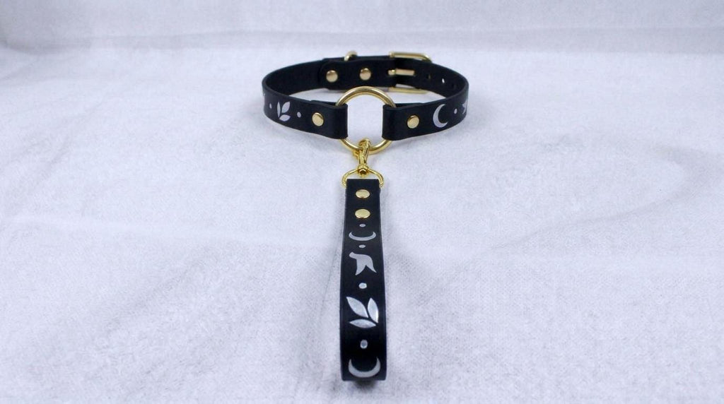 A black leather collar with a gold-colored metal buckle and a dangling black strap with white floral designs.
