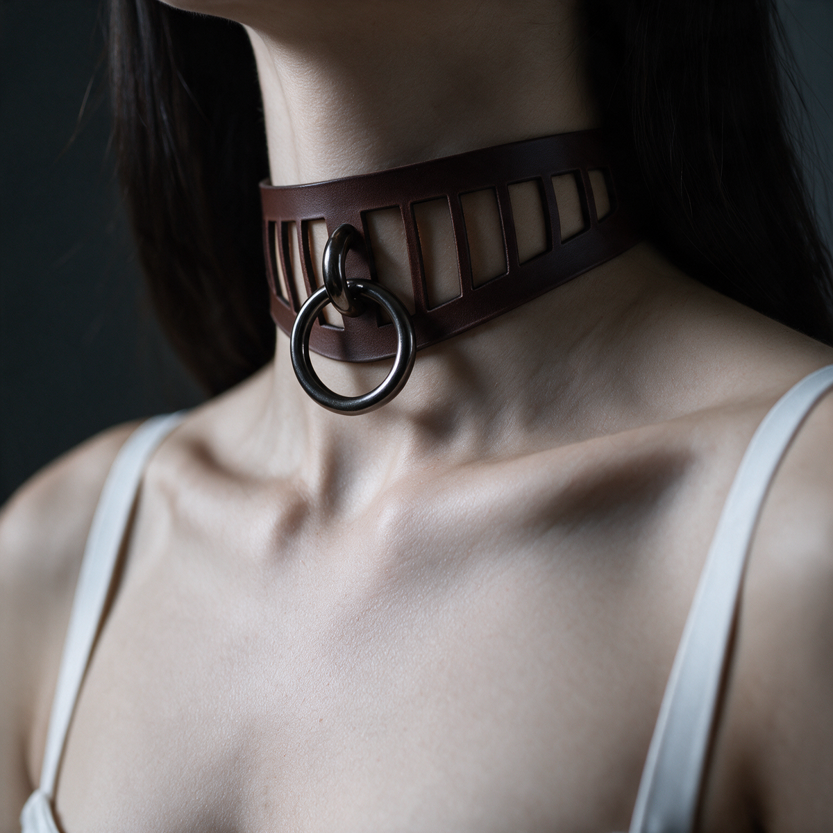A close-up of a woman's neck and chest area wearing a dark leather choker necklace with a metal ring.