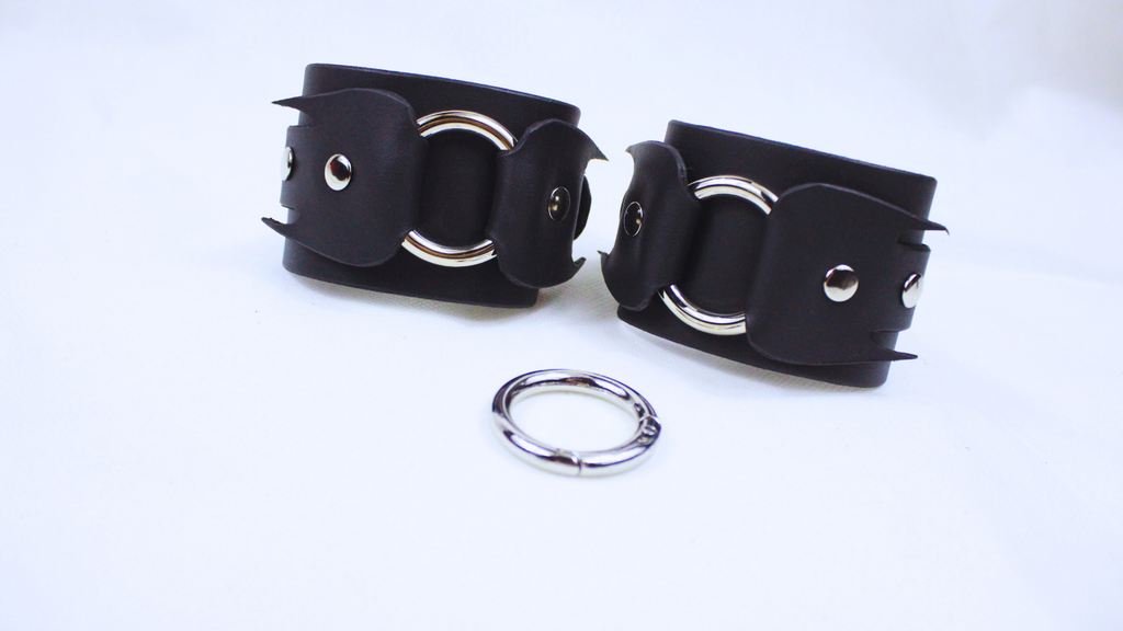a pair of black leather straps with silver rings and a silver ring connector, arranged on a white background.