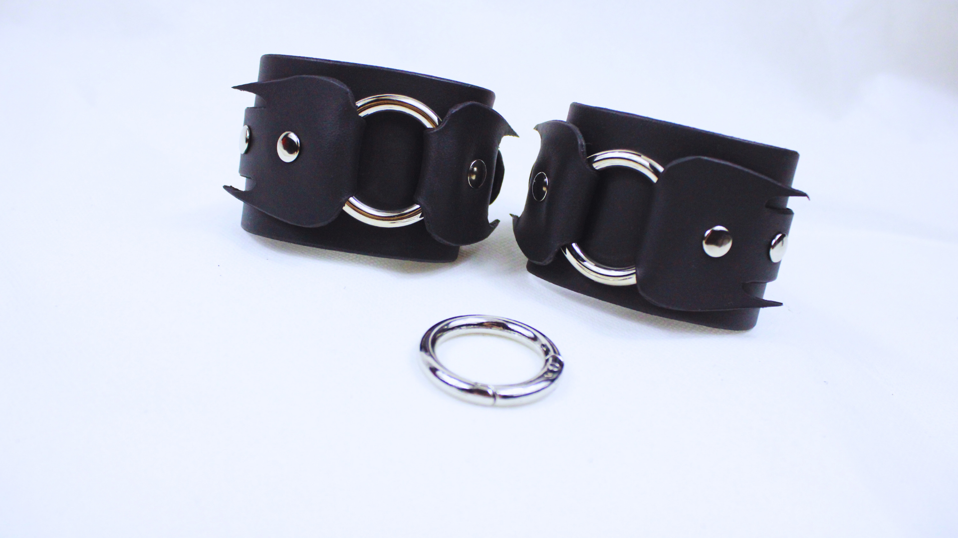 a pair of black leather straps with silver rings and a silver ring connector, arranged on a white background.
