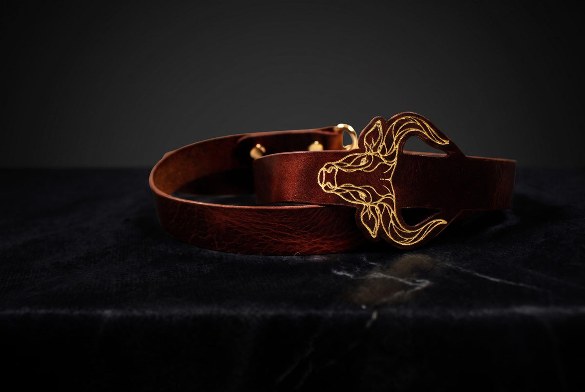A brown leather belt with a gold-colored buckle featuring an animal design, placed on a dark surface.