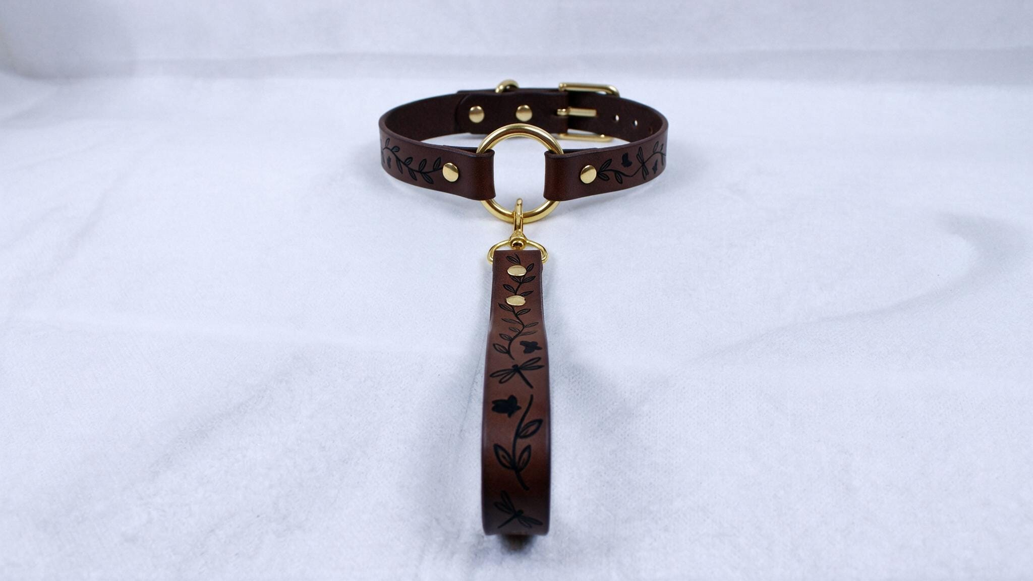 A brown leather collar with a gold buckle and a black handle with a floral design.