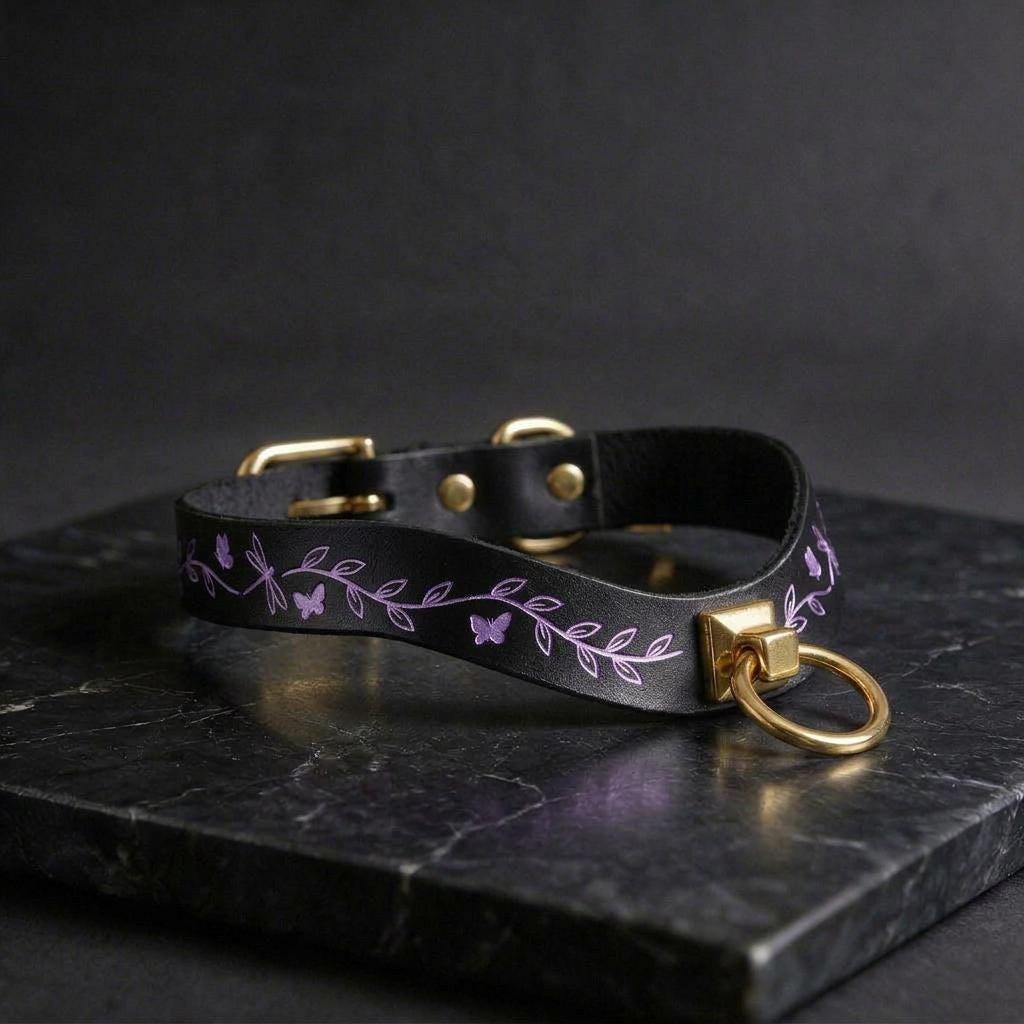 A black leather collar with a gold-colored metal ring and a purple leaf design on it, resting on a black marble surface.