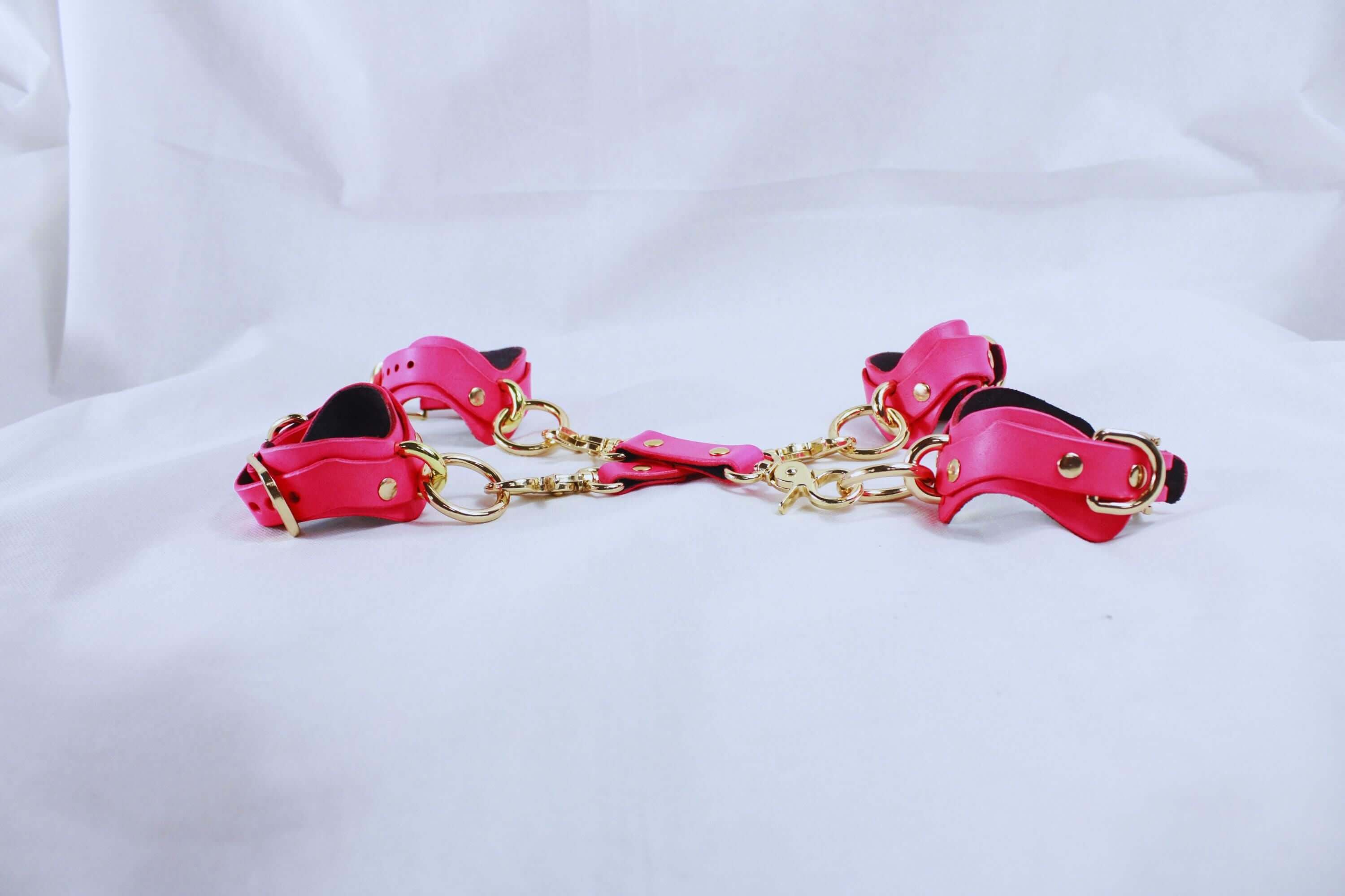 Pink leather BDSM bondage gear set with matching wrist and ankle cuffs connected by golden hardware.