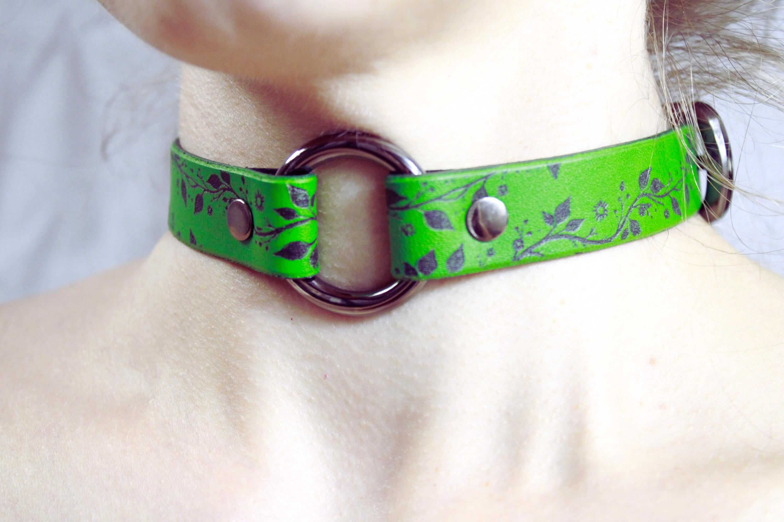 A close-up of a person's neck wearing a green leather choker necklace with a metal ring and two metal buttons.