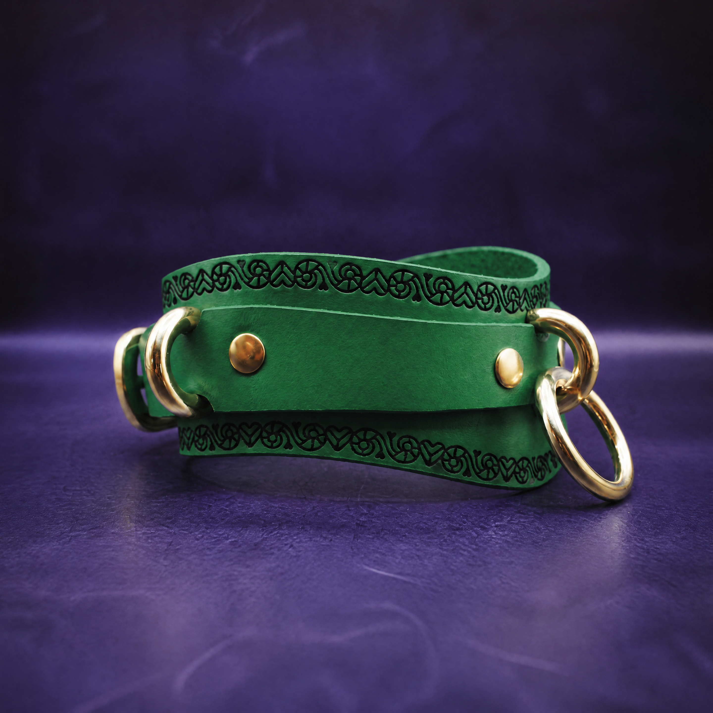 A green leather choker necklace with gold-colored metal rings and a decorative pattern on the front.